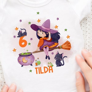 Könnte beinhalten: Ein weißer Strampler mit einer Aquarell-Illustration einer Hexe, die auf einem Besen fliegt. Die Hexe trägt ein lila Kleid und einen schwarzen Hut. Die Illustration enthält Sterne, einen Kessel und zwei schwarze Katzen. Die Zahl 6 und der Name "TILDA" sind ebenfalls enthalten.