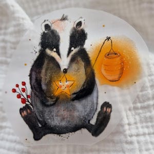 May include: A watercolor illustration of a badger holding a yellow star with a smiling face. The badger is sitting on a white background with a red berry branch and a yellow lantern hanging in the background.