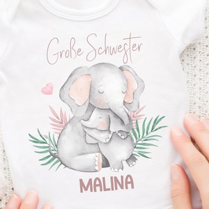 Peut inclure: Combinaison blanche pour bébé avec une illustration aquarelle de deux éléphants qui se font un câlin. Le texte "Große Schwester" et "MALINA" sont imprimés en rose sur la combinaison.