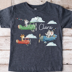 Peut inclure: Un t-shirt bleu foncé avec un motif de trois animaux volant dans des avions. Les animaux sont une girafe, un éléphant et un lapin. Le texte "2 Clara" est imprimé sur le t-shirt.