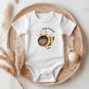 May include: White baby onesie featuring a cartoon bumblebee illustration. The bee is yellow and black with a brown head and red cheeks. The text "Wilde Hummel Lene" is printed below the bee.