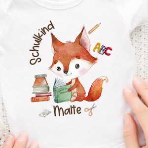 Puede incluir: Un body blanco para bebé con una ilustración de un zorro marrón sosteniendo un libro. El body tiene el texto "Schulkid Malte" y un lápiz, unas tijeras y las letras "ABC" impresas en él.