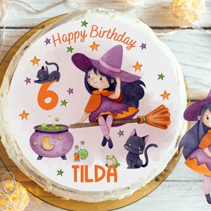 Puede incluir: Un pastel redondo rosa y blanco con una bruja morada y naranja en una escoba, un caldero y dos gatos negros. El pastel está decorado con estrellas y el texto "Happy Birthday" y "Tilda".