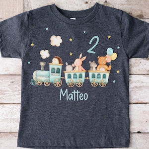 Könnte beinhalten: Ein dunkelgraues T-Shirt mit einem blauen Zug, der einen Igel, ein Kaninchen, eine Maus und einen Bären transportiert. Der Zug hat eine weiße Wolke darüber und gelbe Sterne. Die Zahl 2 befindet sich über dem Zug und der Name Matteo unter dem Zug.
