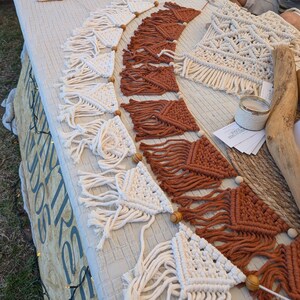 Puede incluir: Exhibición de artesanías de macramé hechas a mano, incluyendo guirnaldas y pequeños tapices de pared. Las guirnaldas presentan diseños triangulares y rectangulares en crema y óxido, con detalles de cuentas de madera. Otros artículos incluyen posavasos y pequeños tapices.