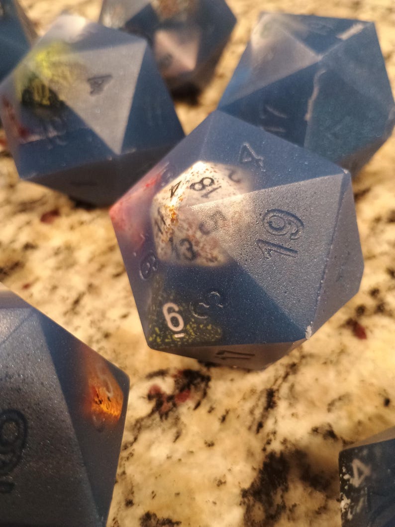 May include: A set of blue, multi-sided dice with white numbers. The dice are made of a textured material and have a speckled appearance. The numbers on the dice are 1, 2, 3, 4, 5, 6, 7, 8, 9, 10, 11, and 12.