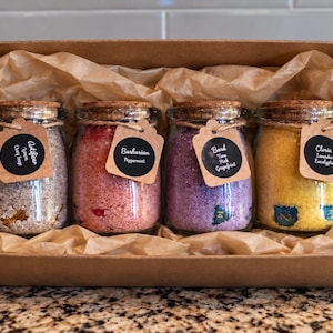 May include: A brown cardboard gift box containing four glass jars with cork lids, filled with colorful bath salts. Each jar has a black circular tag with white text: Artificer, Barbarian, Bard, and Cleric, along with scent and class labels.