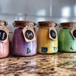 Può includere: Una fila di barattoli di vetro riempiti con sali da bagno colorati, ciascuno sormontato da un tappo di sughero e una piccola etichetta di carta marrone legata con dello spago. I sali variano nei colori rosa, viola, giallo, verde e blu, con inclusioni botaniche.