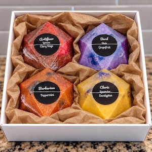 Könnte beinhalten: Eine weiße Geschenkbox mit vier geometrischen, polyederförmigen Seifen, die in Klarsichtfolie eingewickelt und in braunes Knitterpapier gebettet sind. Jede Seife hat ein schwarzes, rundes Etikett mit weißem Text: Artificer Lemon Clary Sage, Bard Pink Grapefruit, Barbarian Peppermint und Cleric Lavender Eucalyptus.