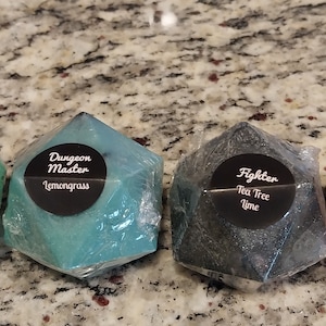 May include: Four green, blue, black, and brown geometric soaps shaped like a 20-sided die. Each soap has a black circle label with white text. The labels read "Druid Clary Sage Rosemary", "Dungeon Master Lemongrass", "Fighter Tea Tree Lime", and "Monk Tea Tree Bergamot".