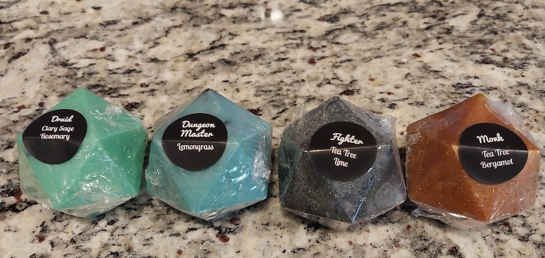 May include: Four green, blue, black, and brown geometric soaps shaped like a 20-sided die. Each soap has a black circle label with white text. The labels read "Druid Clary Sage Rosemary", "Dungeon Master Lemongrass", "Fighter Tea Tree Lime", and "Monk Tea Tree Bergamot".