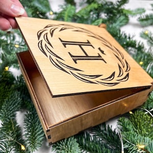 May include: A wooden box with a laser-etched design on the lid. The lid features a wreath design around the letter "H". The box is open, revealing the interior. The box is made of light-colored wood and is surrounded by greenery.