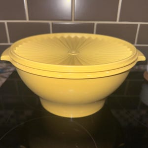 May include: A bright yellow plastic mixing bowl with a matching lid. The lid features a distinct sunburst pattern of radiating lines extending from the center. The bowl has a wide, rounded body that tapers down to a circular base and includes side handles.