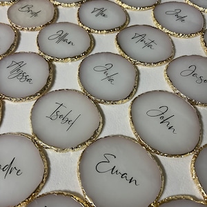 May include: A set of round, white coasters with gold trim and black calligraphy names. The names include Alyssa, Isobel, Ewan, John, Julia, Anita, Rose, Bryant, Hugh, Ruby, Groom, Bride, Jillian, Alex, Jodie, and Jason.