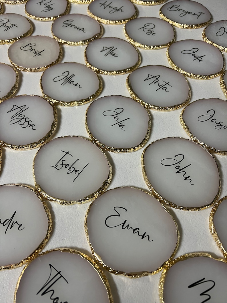 May include: A set of round, white coasters with gold trim and black calligraphy names. The names include Alyssa, Isobel, Ewan, John, Julia, Anita, Rose, Bryant, Hugh, Ruby, Groom, Bride, Jillian, Alex, Jodie, and Jason.