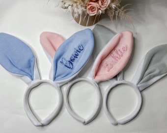 Peut inclure: Trois bandeaux d'oreilles de lapin avec des bandes blanches et des oreilles de différentes couleurs. Les oreilles sont bleues avec le nom "Berrie" brodé en paillettes bleues, roses avec le nom "Zalie" brodé en paillettes roses et grises.
