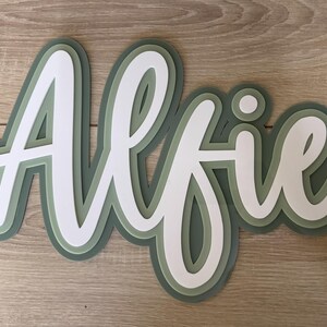 May include: A layered wooden sign spelling the name "Alfie" in a cursive font. The sign has a white top layer, a light green middle layer, and a darker green base layer. The sign is placed on a light brown wooden surface.
