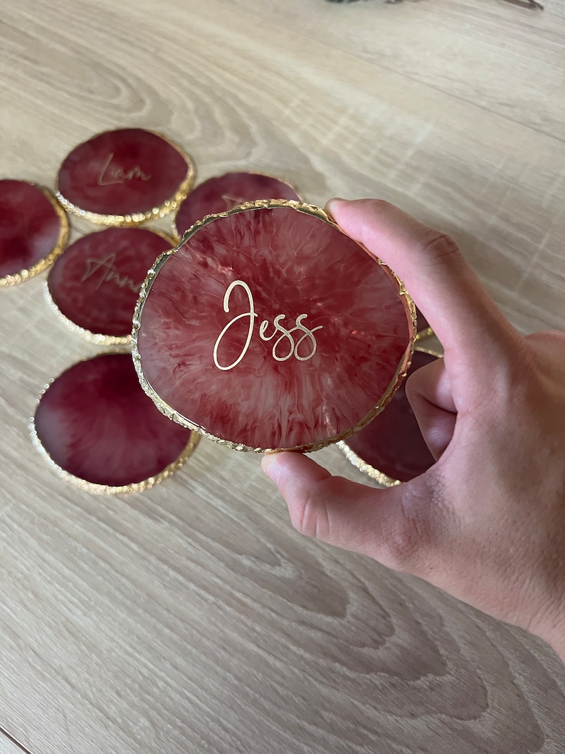May include: Set of six red and gold resin coasters with gold foil lettering. The coasters feature the names "Liam", "Ann", "Jess", and three other names.