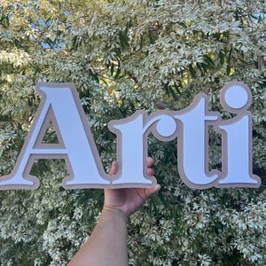May include: White letters spelling "Arti" with a beige outline. The letters are raised and have a 3D effect.