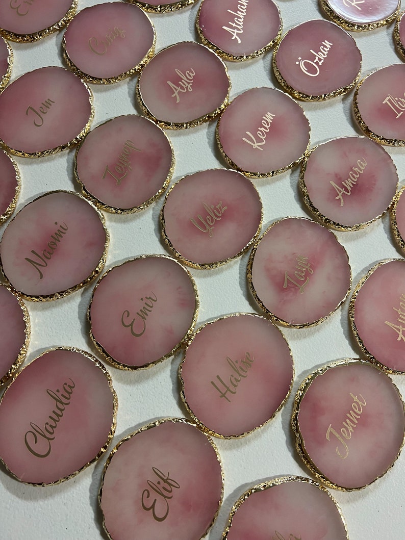 May include: Pink and gold round coasters with gold trim and names written in gold script. The names include Jem, Naomi, Temen, Ayla, Atahar, Claudia, Emir, Ueliz, Kerem, &Ouml;zkan, Elif, Halise, Tagu, Amara, Ilix, and Jennet.