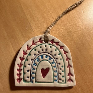May include: A white ceramic ornament shaped like a rainbow with a red heart in the centre. The rainbow is decorated with red, green, blue, and purple details. The ornament is attached to a brown twine string.
