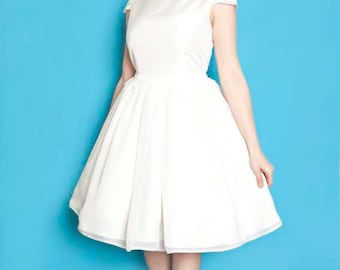 Puede incluir: Una mujer con un vestido blanco hasta la rodilla con ribete de encaje y escote barco. Tiene el pelo rojo y lleva pendientes blancos. Está de pie frente a un fondo azul.