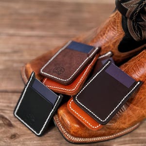 Puede incluir: Cuatro porta tarjetas de cuero en marrón, negro y naranja con costuras blancas. Los porta tarjetas están apilados uno encima del otro y están sobre una superficie de madera.