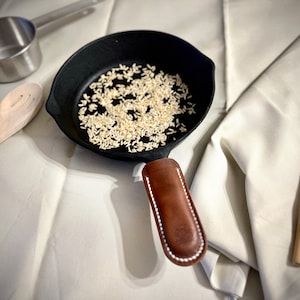 May include: A black cast iron skillet with a brown leather handle, filled with uncooked white rice. The skillet is sitting on a white surface next to a wooden spoon.