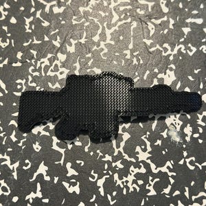 May include: A black, textured, gun-shaped object. The object has a detailed, patterned surface and is set against a background with a white and gray pattern.