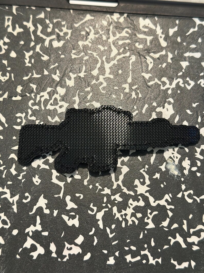 May include: A black, textured, gun-shaped object. The object has a detailed, patterned surface and is set against a background with a white and gray pattern.