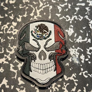 May include: A skull-shaped patch featuring the Mexican flag's colors: green, white, and red. The skull's design includes a detailed white jawline and eye sockets, with the Mexican coat of arms in the center. The patch has a black border.