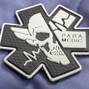 K&ouml;nnte beinhalten: Ein schwarz-wei&szlig;es Sanit&auml;terabzeichen in Form eines Kreuzes. Das Abzeichen zeigt einen Totenkopf mit Fl&uuml;geln und das Wort "PARAMEDIC". Der Hintergrund des Abzeichens hat ein strukturiertes Muster.