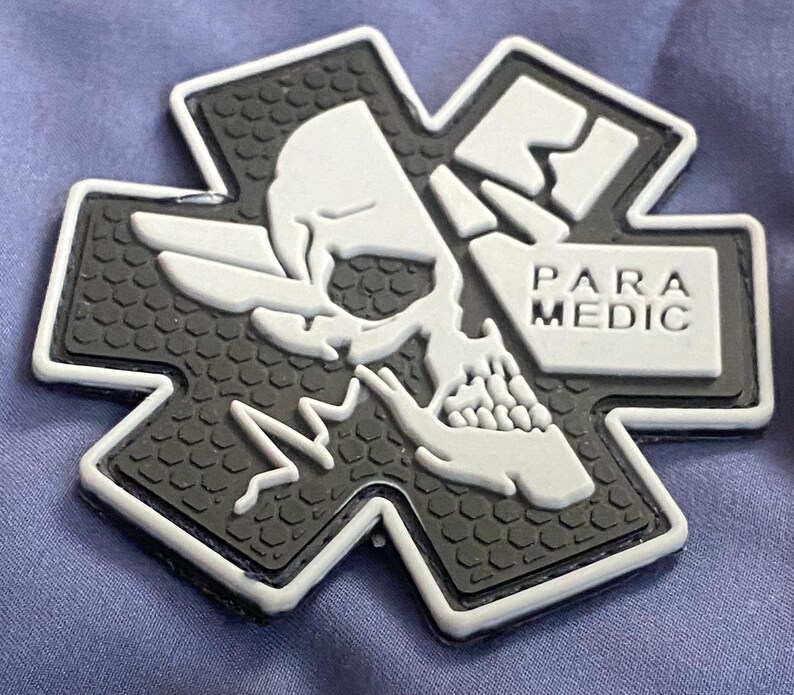 K&ouml;nnte beinhalten: Ein schwarz-wei&szlig;es Sanit&auml;terabzeichen in Form eines Kreuzes. Das Abzeichen zeigt einen Totenkopf mit Fl&uuml;geln und das Wort "PARAMEDIC". Der Hintergrund des Abzeichens hat ein strukturiertes Muster.