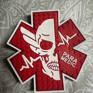 May include: A red and white Star of Life emblem featuring a skull design on one side and the words "PARA MEDIC" on the other. The emblem has a textured surface and a white border.