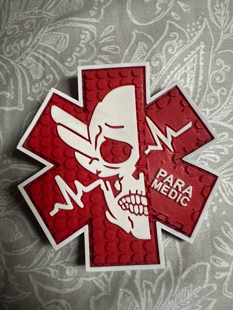 May include: A red and white Star of Life emblem featuring a skull design on one side and the words "PARA MEDIC" on the other. The emblem has a textured surface and a white border.
