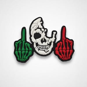 May include: Embroidered patch featuring a white skull with a black outline, flanked by two hands giving the middle finger. One hand is green, the other is red, both with black outlines. The patch has a black border and is set against a white background.