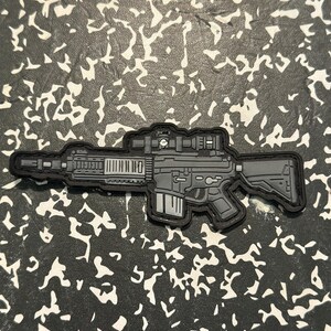 May include: A gray and black patch in the shape of a rifle with a scope. The patch has a detailed design, including the barrel, magazine, and stock. The patch is on a black and white patterned background.