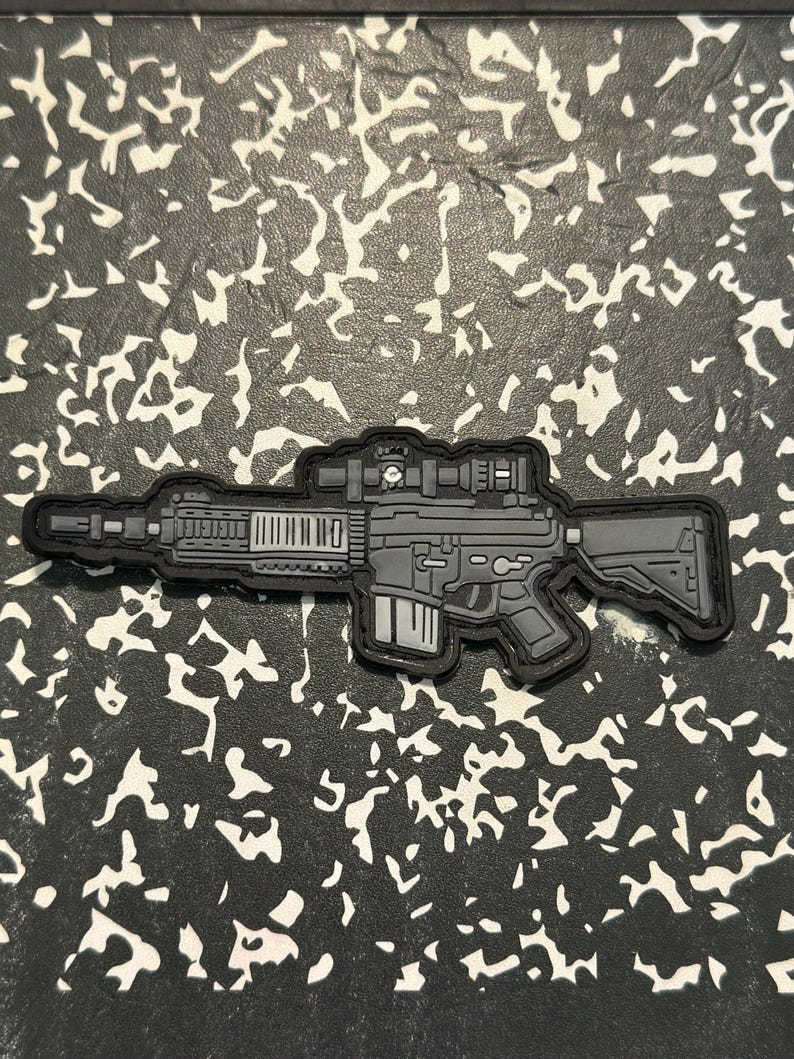 May include: A gray and black patch in the shape of a rifle with a scope. The patch has a detailed design, including the barrel, magazine, and stock. The patch is on a black and white patterned background.
