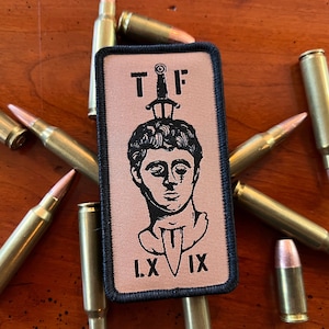 May include: A tan and black embroidered patch with a black and white illustration of a person with a sword through their head. The text "TIF" is above the person and "L.X.IX" is below the person.
