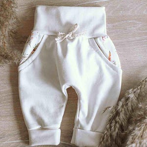 Könnte beinhalten: Cremeweiße Hose mit hohem Bund und einem dekorativen Spitzen-Kordelzug. Die Hose verfügt über zwei Seitentaschen, die mit einem zarten Blumenmuster in Pastelltönen gefüttert sind. Das Kleidungsstück hat Beinbündchen und eine lockere Passform.