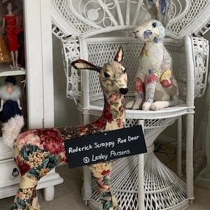 May include: A white wicker peacock chair with a floral patterned fabric roe deer and a floral patterned fabric bunny sitting on the seat. A black chalkboard sign reads "Roderick Scrappy Roe Deer © Lesley Parsons".