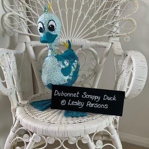 May include: A blue and white patchwork fabric duck sitting on a white wicker chair. The duck has a teal beak and large black eyes. A black sign reads "Dubonnet Scrappy Duck © Lesley Parsons".