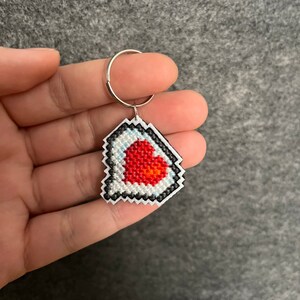 May include: A white and black heart-shaped keychain with a red heart embroidered in the center. The keychain is attached to a silver ring.