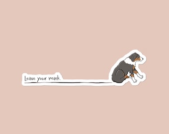 Könnte beinhalten: Ein Aufkleber mit einem Cartoon-Hund in Grau-, Weiß- und Brauntönen, der sich kratzt. Der Text "Leave your mark" befindet sich über einer gezogenen Linie, die sich vom Hund erstreckt. Der Hintergrund ist einfarbiges, gedämpftes Rosa.