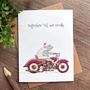 May include: A greeting card with a cartoon illustration of two frogs riding a red motorcycle. The card has the text "Together 'til we croak" in black script. A small green plant and a pencil are in the background.