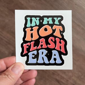 May include: A square sticker with the text "IN MY HOT FLASH ERA" in a retro font. The text is in shades of blue, orange, and red with a black outline. The sticker is held by a hand.