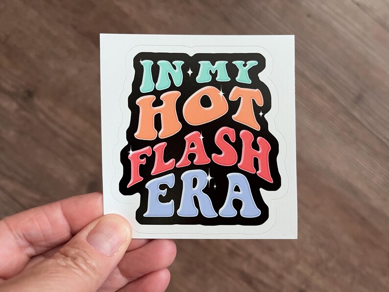 May include: A square sticker with the text "IN MY HOT FLASH ERA" in a retro font. The text is in shades of blue, orange, and red with a black outline. The sticker is held by a hand.