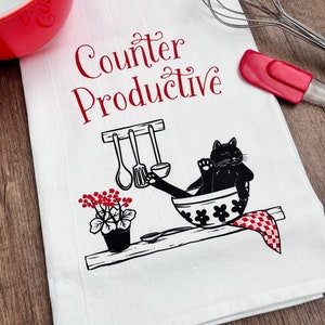 May include: White kitchen towel with red text that says "Counter Productive" and a black cat sitting in a white bowl with red flowers. The cat is holding a spoon in its paw.