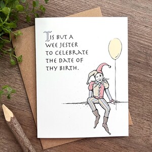 May include: A white card with a black and white illustration of a jester holding a yellow balloon. The card has the text "'Tis but a wee jester to celebrate the date of thy birth."