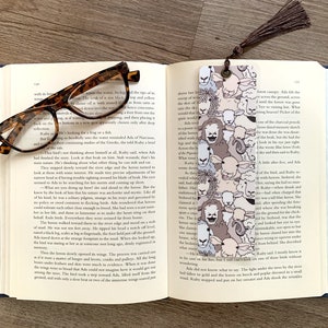 May include: A close-up of a bookmark with a pattern of sheep. The bookmark is made of a clear material and has a brown tassel. The bookmark is lying on top of an open book.